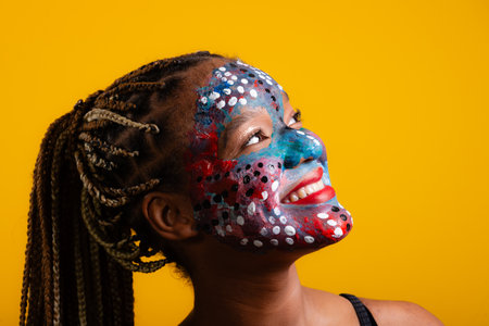 Close-up portrait of beautiful young woman's face with braids in her hair and face painted with colorful paint. Isolated on yellow background.の写真素材