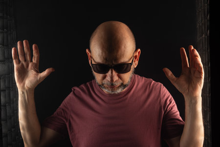 Bald and bearded man wearing sunglasses with hands raised. Isolated on dark smoky background.
