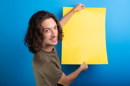 smiling young black haired man holding empty yellow poster looking at camera against blue...