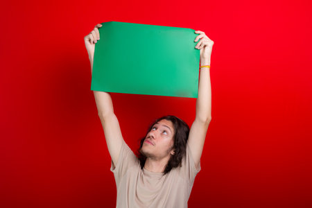 Young handsome caucasian man holding green colored, empty poster with arms up isolated on red...