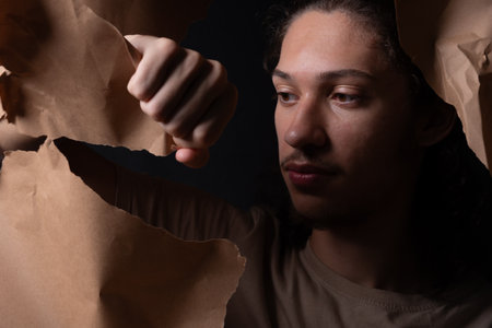 A young man coming out through a hole in a sheet of brown paper. closeup. Studio portrait.の写真素材