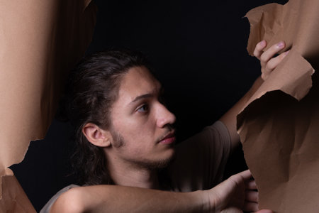 A man peering through a hole in a sheet of brown paper. Tearing the paper with his hands. Studio portrait.の写真素材
