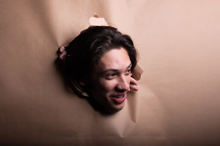 Man's head sticking out and looking through a paper hole with his hands pushing the paper aside. Studio portrait.の写真素材