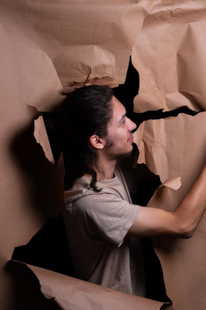 A man peering through a hole in a sheet of brown paper. Tearing the paper with his hands. Studio portrait.の写真素材