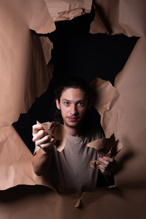 A man coming out through a hole in a sheet of brown paper. Tearing with and showing with his hands the paper. Studio portrait.の写真素材