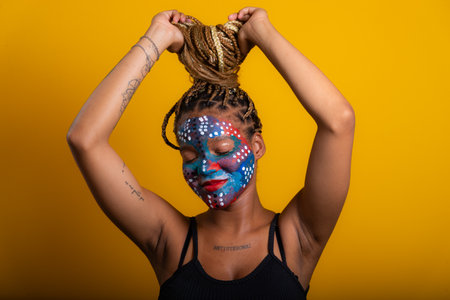 portrait of beautiful young woman with colorful paint painted face touching braids in hair with closed eyes. Isolated on yellow background.の写真素材