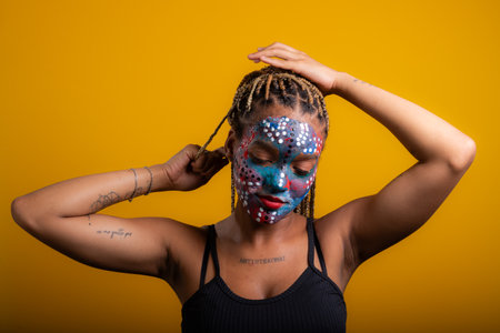 Young woman wearing black blouse holding braids in her hair with face painted with colorful paint, concentrated woman. Isolated on yellow background.の写真素材