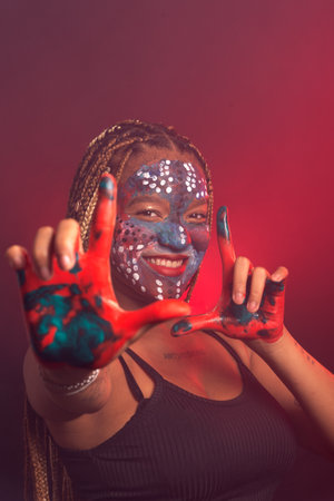 Woman with face and hands painted with colorful paint and braids in her hair making gestures with her hands and fingers. Isolated on a background with red smoke.の写真素材