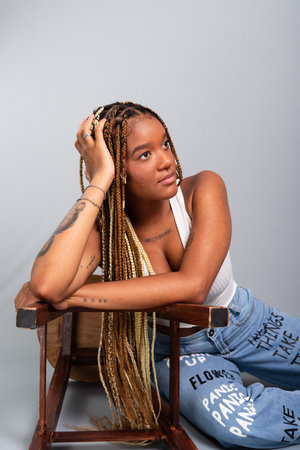 Beautiful brunette woman with braids in her hair, sitting on the studio floor leaning on a wooden stool. Calm and confident person. Isolated on gray background.の写真素材