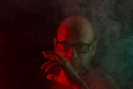 Close-up portrait of bald and bearded man in prescription glasses posing for photo. Isolated on dark smoky background with colored lights. South American person.の写真素材