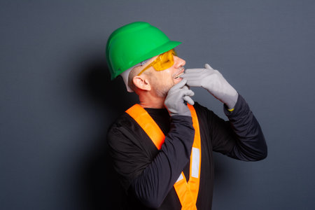 Photo of a civilian worker wearing a green helmet, reflective vest and protective glasses, biting his glove. Isolated on gray background. Construction worker.の写真素材