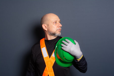 Photo of a civilian worker in gloves and reflective vest holding green helmet against his chest. Isolated on gray background. Construction worker.の写真素材