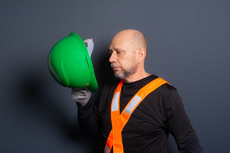 Photo of a civilian worker in gloves and reflective vest holding green helmet against his chest. Isolated on gray background. Construction worker.の写真素材