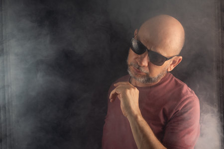 Bald and bearded man wearing sunglasses posing for a photo in a studio with a smoky black background.の写真素材