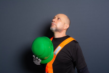 Photo of a civilian worker in gloves and reflective vest holding green helmet against his chest. Isolated on gray background. Construction worker.の写真素材
