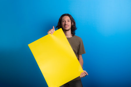 young man holding a yellow, empty poster, looking at camera against blue background. advertisement concept.の写真素材