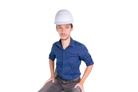 Studio portrait of young architect sitting wearing white helmet and looking at camera. Isolated on...
