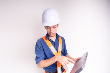Young architect wearing white helmet and luminous vest, working with laptop. Isolated on white...