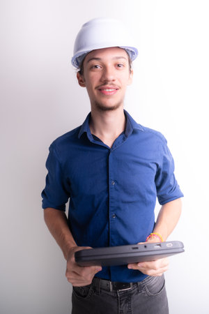 Young architect wearing white helmet and luminous vest, holding a laptop and looking at the camera. Isolated on white background. Safety equipment.の写真素材