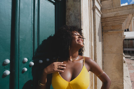 Beautiful black woman with raven hair wearing yellow dress posing for photo against big green church...