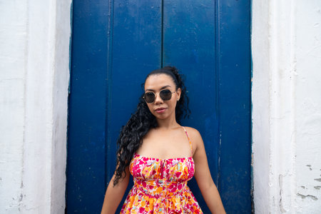 Beautiful young woman with braids and colorful dress sitting in front of a blue door of an old house...