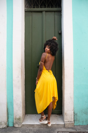 Portrait of beautiful black woman with afro hair wearing yellow dress posing for photo leaning against a green door. Happy person on trip.の写真素材