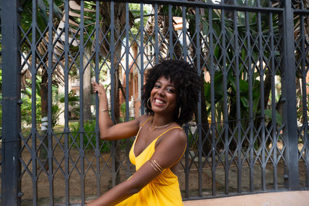 Happy smiling woman with afro hair wearing yellow dress posing for the camera leaning against an old iron fence. Happy person on a trip.の写真素材