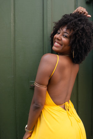 Portrait of beautiful black woman with afro hair wearing yellow dress posing for photo leaning against a green door. Happy person on trip.の写真素材