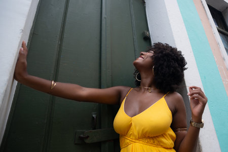 Portrait of beautiful black woman with afro hair wearing yellow dress posing for photo leaning against a green door. Happy person on trip.の写真素材