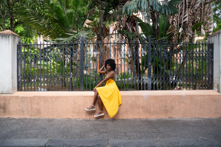 Happy smiling woman with afro hair wearing a yellow dress sitting next to an old iron fence. Happy person on a trip.の写真素材