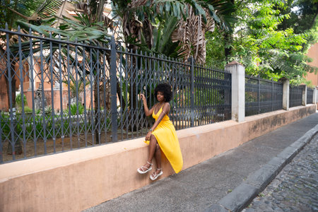 Happy smiling woman with afro hair wearing a yellow dress sitting next to an old iron fence. Happy person on a trip.の写真素材