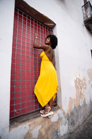 Woman wearing yellow clothes with afro hair holding an iron railing with a red door looking to the side. Confident and happy personの写真素材
