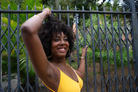 Portrait of a beautiful black woman with afro hair wearing a yellow dress leaning against an old iron fence. Happy person on a trip.の写真素材