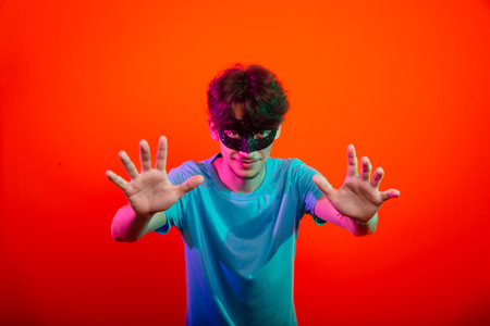 Handsome young man wearing carnival mask looking and making hand gestures to the camera. Isolated on orange neon background.の写真素材