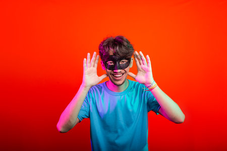 Happy handsome young man wearing carnival mask looking and making hand gestures to the camera. Isolated on orange neon background. Brazilの写真素材
