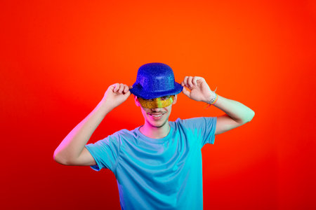 Happy and fun young man in blue shirt, wearing carnival mask and holding blue hat. Posing for the camera. Isolated on neon orange background. Ready for party.の写真素材