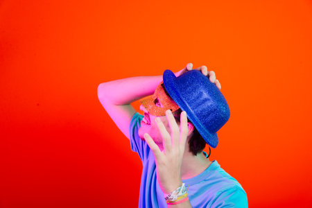 Young man in blue shirt, wearing carnival mask and blue hat looking up. Isolated on neon orange background. Happy and fun person.の写真素材