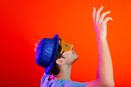 Young man in blue shirt, wearing carnival mask and blue hat looking up. Isolated on neon orange background. Happy and fun person.の写真素材