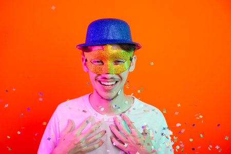 Young man wearing carnival mask and blue hat looking at camera. Confetti flying. Isolated on neon orange background. Happy and fun person.の写真素材