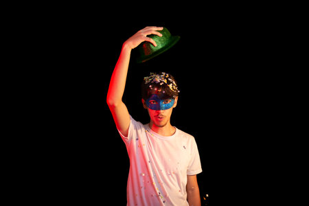 Young man in white shirt wearing carnival mask having fun with hat and paper confetti. Isolated on black background. Happy and cheerful person.の写真素材
