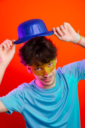 Happy and fun young man in blue shirt, wearing carnival mask and holding blue hat. Posing for the camera. Isolated on neon orange background. Ready for party.の写真素材