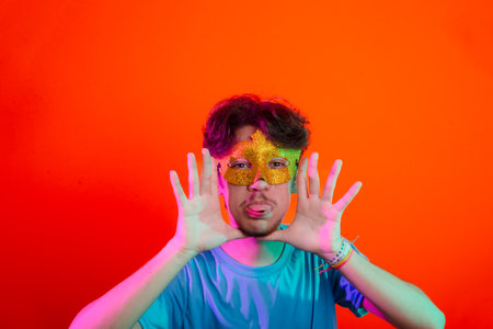 Happy handsome young man wearing carnival mask looking and making hand gestures to the camera. Isolated on orange neon background.の写真素材