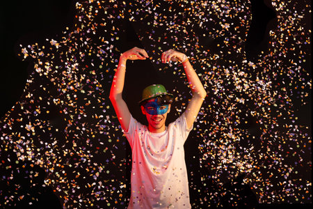 Young man in white shirt, wearing carnival mask and green hat having fun with paper confetti. Isolated on black background. Happy and cheerful person.の写真素材