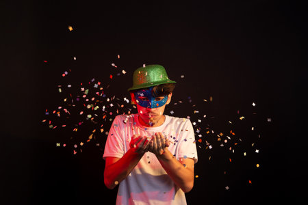 Young man in white shirt, wearing carnival mask and green hat having fun with paper confetti. Isolated on black background. Happy and cheerful person.の写真素材