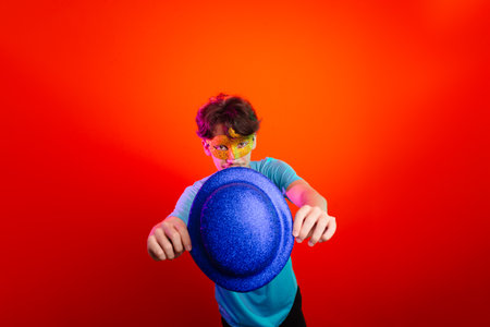 Young man in blue shirt, wearing carnival mask and holding blue hat. Posing for the camera. Isolated on neon orange background. Happy and fun person.の写真素材