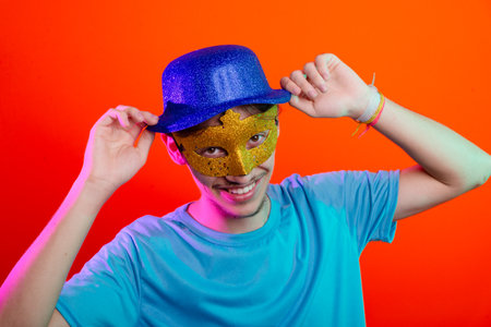 Happy and fun young man in blue shirt, wearing carnival mask and holding blue hat. Posing for the...