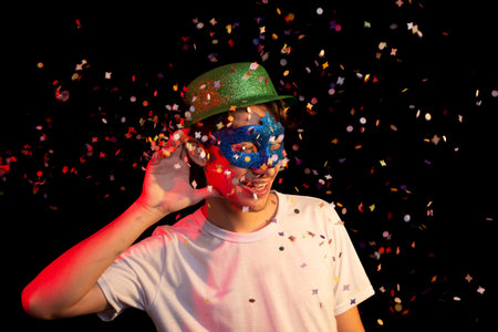 Young man in white shirt, wearing carnival mask and green hat having fun with paper confetti. Isolated on black background. Happy and cheerful person.の写真素材