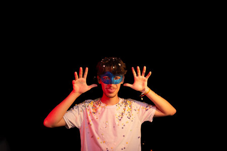 Young man in white shirt, wearing blue carnival mask making hand gestures to camera and having fun with confetti. Isolated on black background. Happy and cheerful person.の写真素材