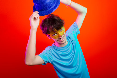 Happy and fun young man in blue shirt, wearing carnival mask and holding blue hat. Posing for the camera. Isolated on neon orange background. Ready for party.の写真素材