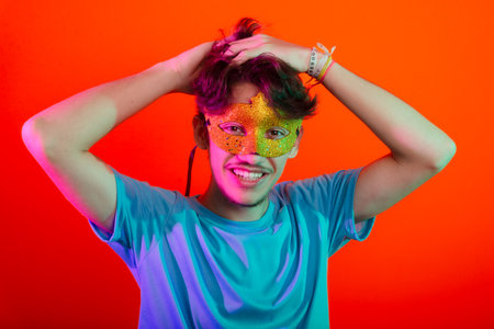 Happy and fun young man wearing carnival mask, touching with both hands his hair. Isolated on neon orange background.の写真素材